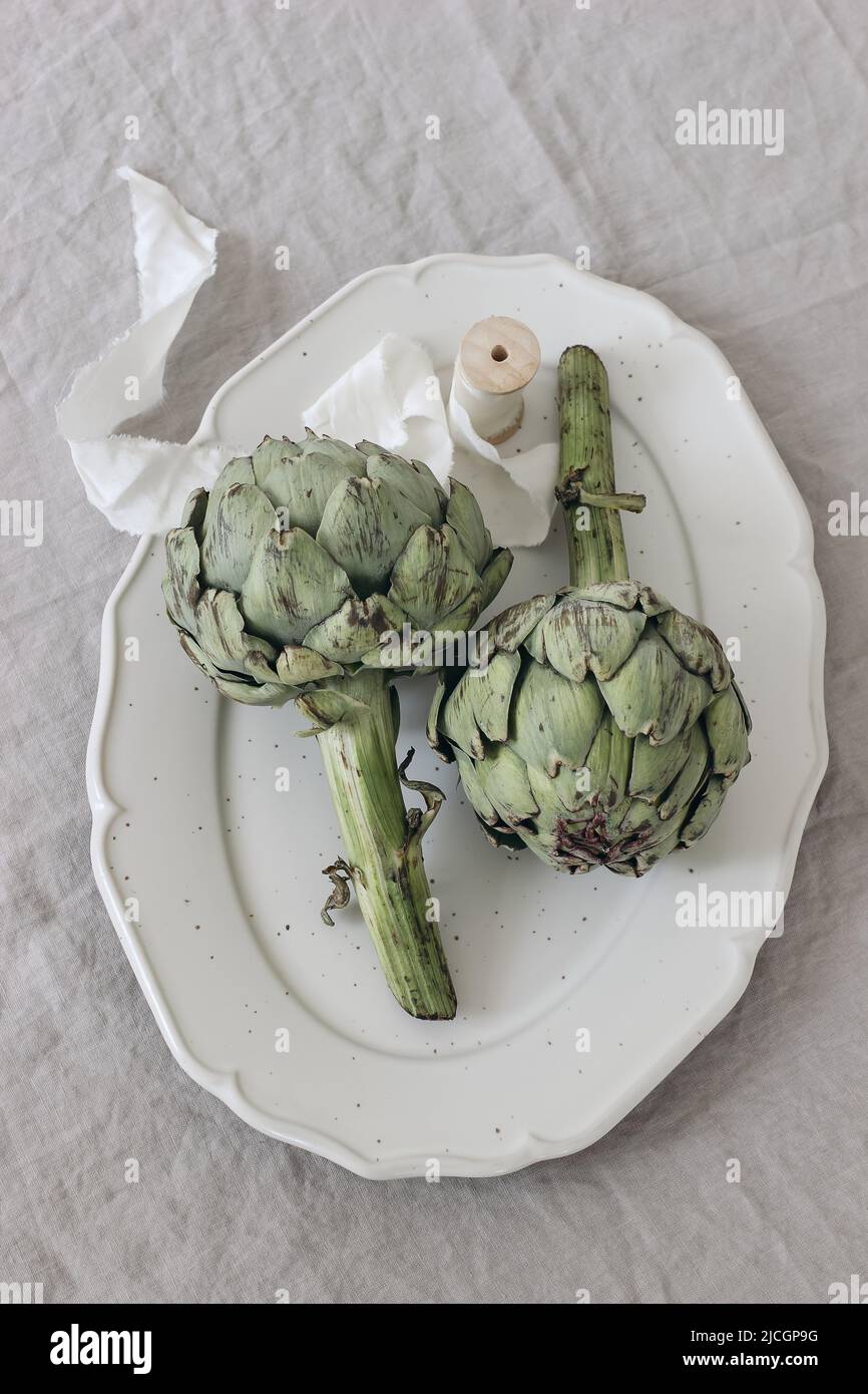 Elegant vegetable still life. Two artichokes and white silk ribbon on porcelain plate, tray. Grey linen table cloth. Feminine farm, rustic lifestyle Stock Photo