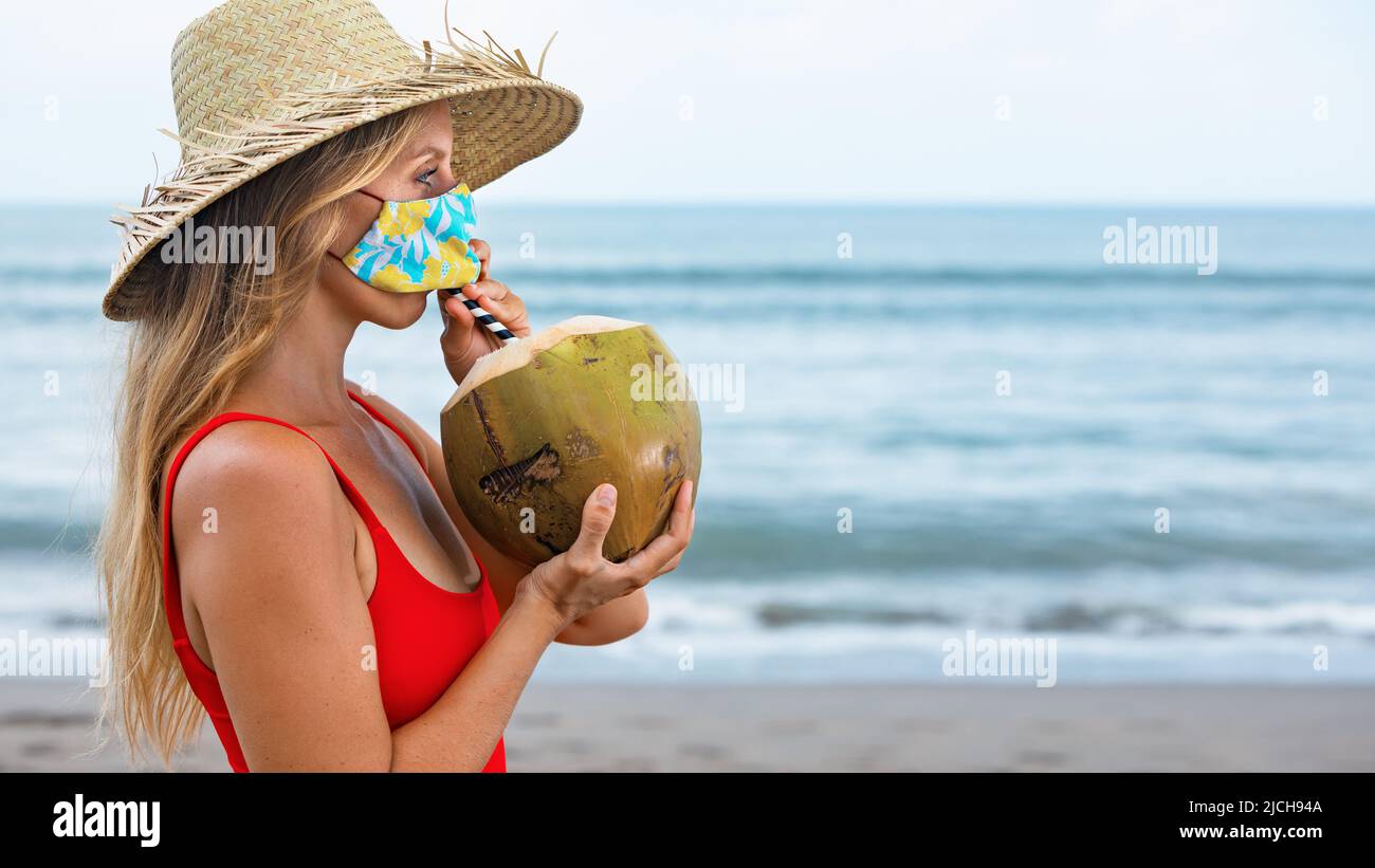 Funny portrait of woman in straw hat drinking young coconut on tropical sea beach. New rules to wear cloth face covering mask at public places Stock Photo