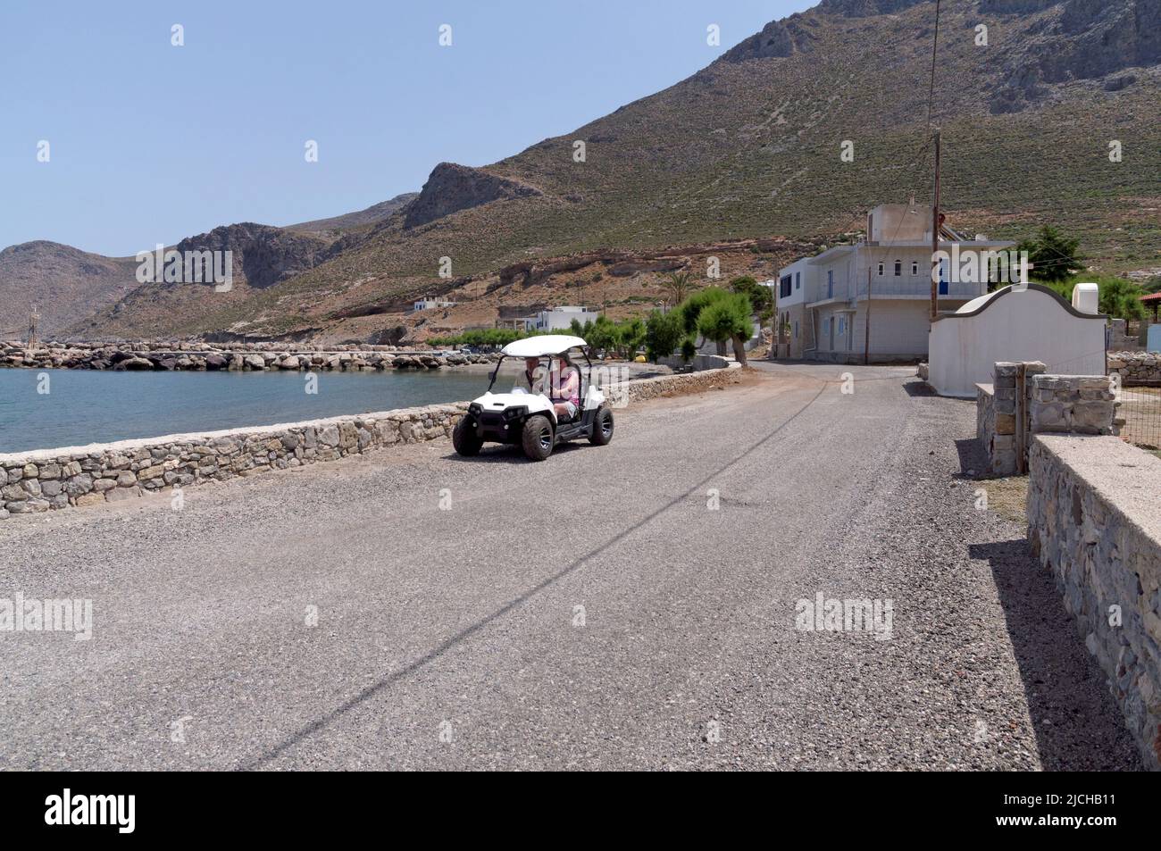 Couple travelling in a Beach buggy on a road by the sea past a small church.. Tilos island Dodecanese, near Rhodes Greece. May 2022. Stock Photo