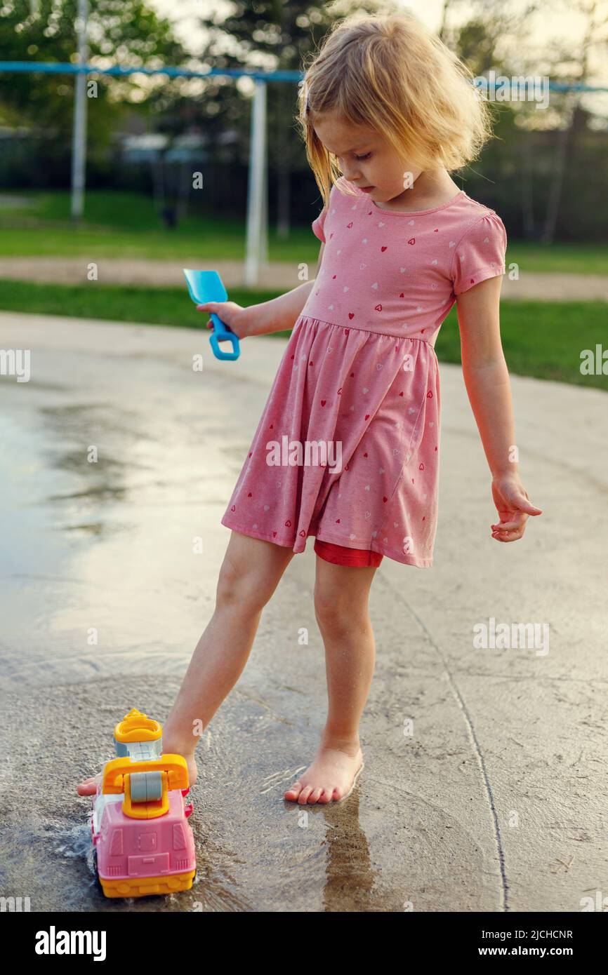 Small child playing at splash pad with water and outdoor toys. Little girl having fun at playground with fountains in summer. Stock Photo