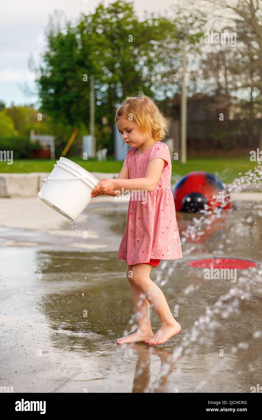 Child playing with bucket at splash pad playground in water park. Girl with fountain in summer. Stock Photo