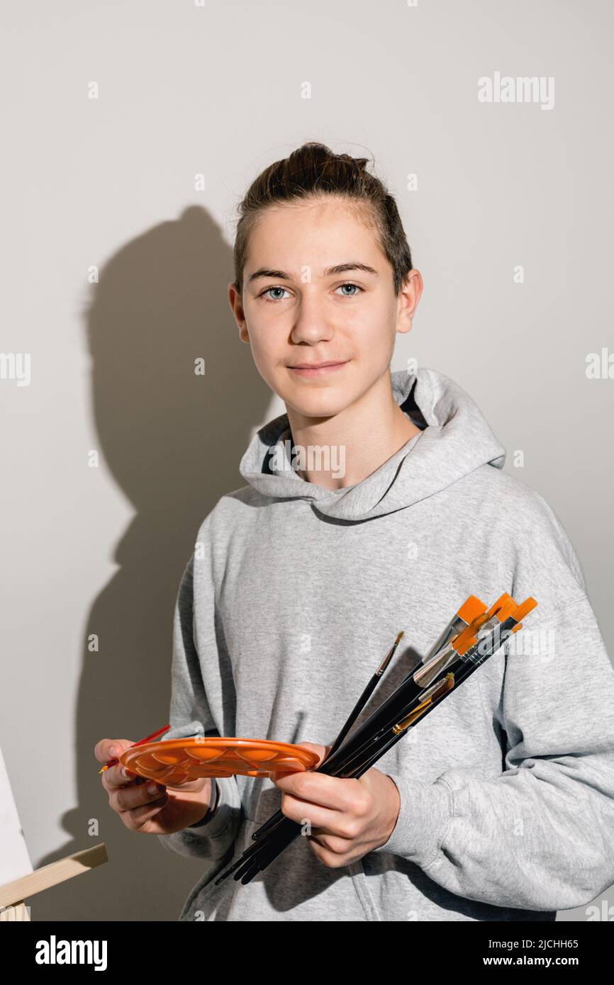 Portrait of a teenage artist holding an orange palette and many brushes in his hands on a background of white wall in the studio. Direct view, sharp light. The guy in casual clothes. Copy space. Stock Photo