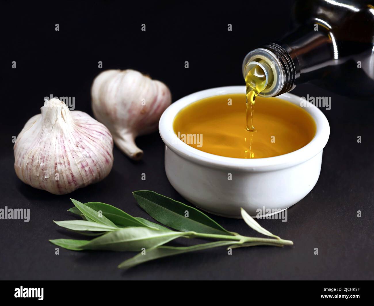 Bottle pouring virgin olive oil in a white bowl on black background, homemade garlic olive oil production Stock Photo