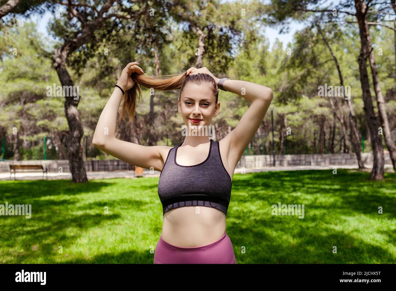 Happy brunette woman wearing black sports bra standing on city park, outdoors tying her hair in a ponytail while looking at the camera. Stock Photo