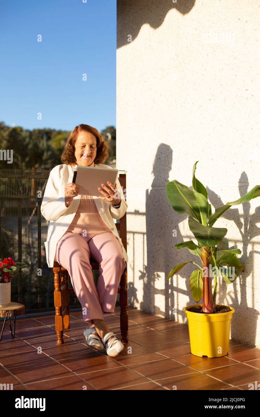 Elderly Caucasian woman using a portable digital tablet on the terrace of her house. Concept of technology, communications and elderly people. Space f Stock Photo