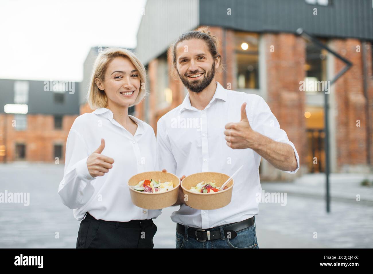 Young cheerful couple, colleague or business partners in white shirts having break and eating healthy meal walking at the city street showing thumb up. Organic healthy nutrition concept. Stock Photo