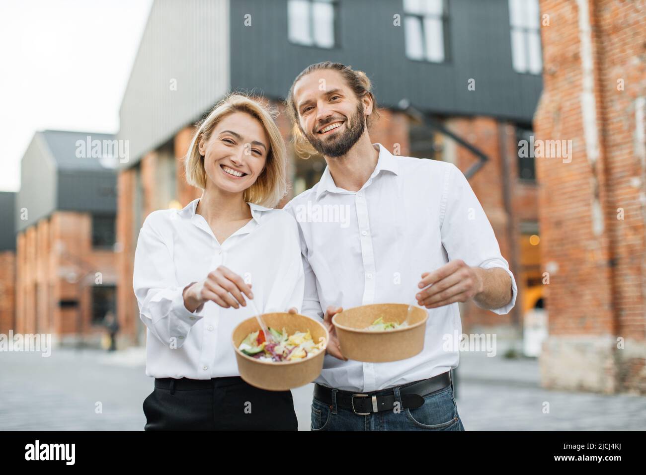 Young cheerful couple, colleague or business partners in white shirts having break and eating healthy meal walking at the city street. Organic healthy nutrition concept. Stock Photo