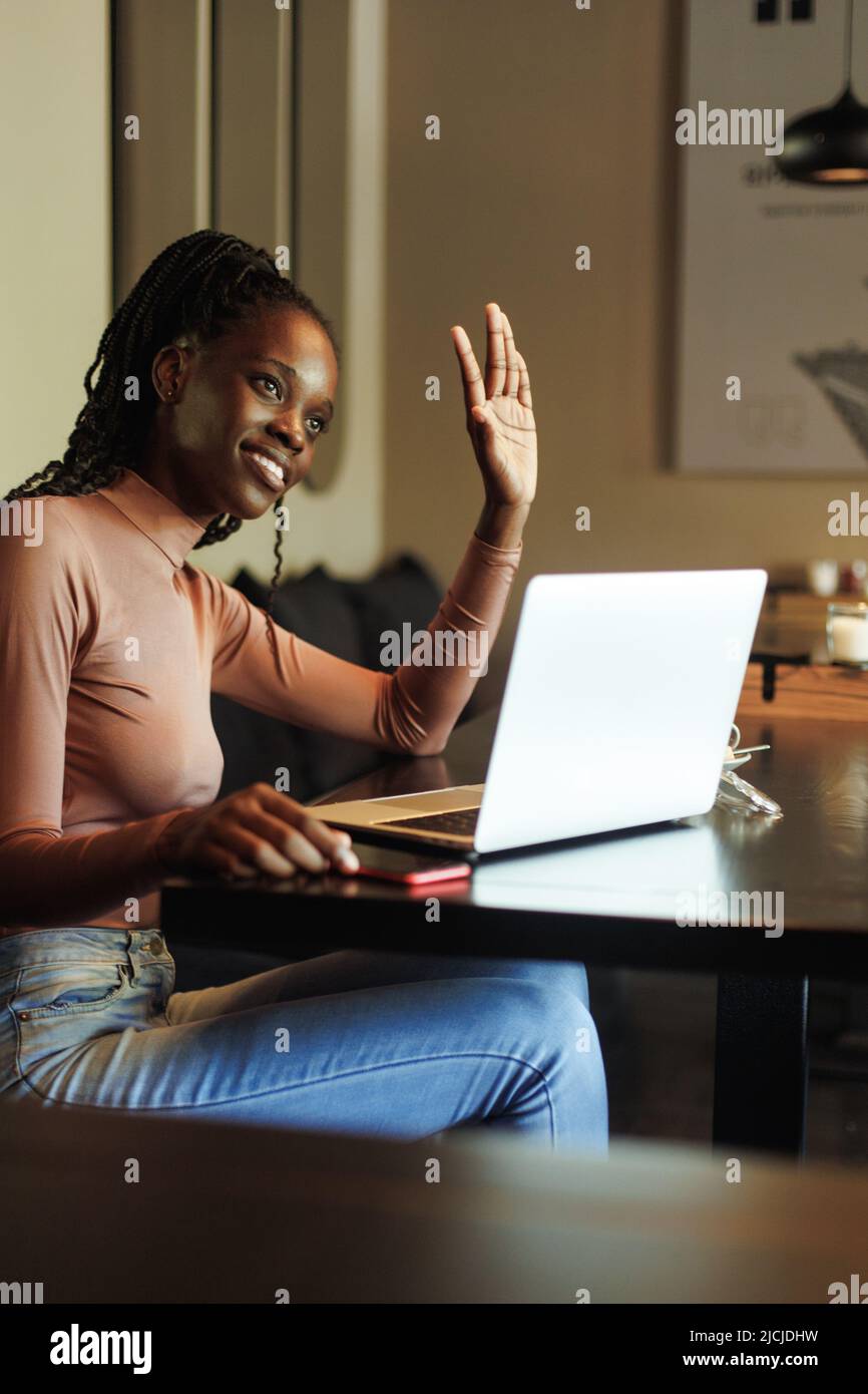 Vertical smiling multiethnic afro american business woman in casual with laptop and smartphone in cafe. Raising hand hi Stock Photo