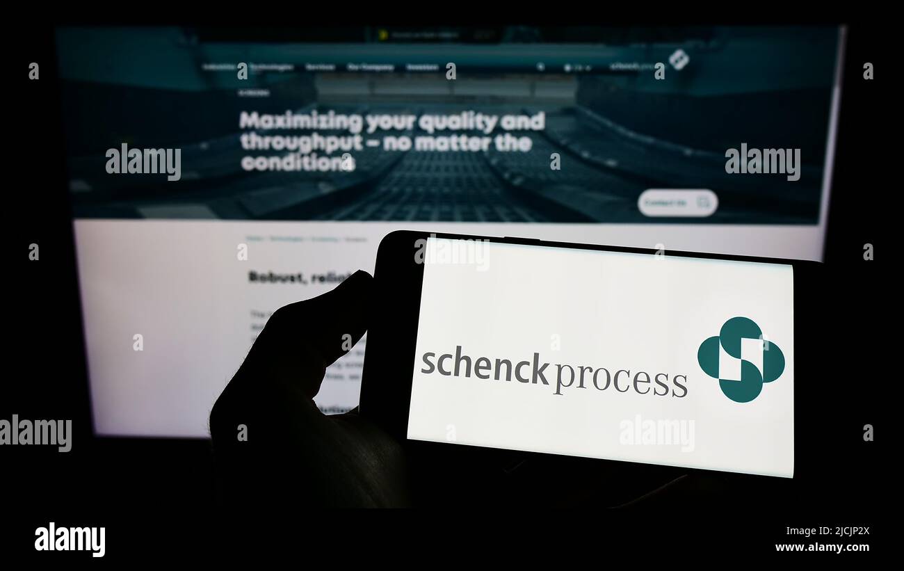 Person holding cellphone with logo of German company Schenck Process Europe GmbH on screen in front of business webpage. Focus on phone display. Stock Photo