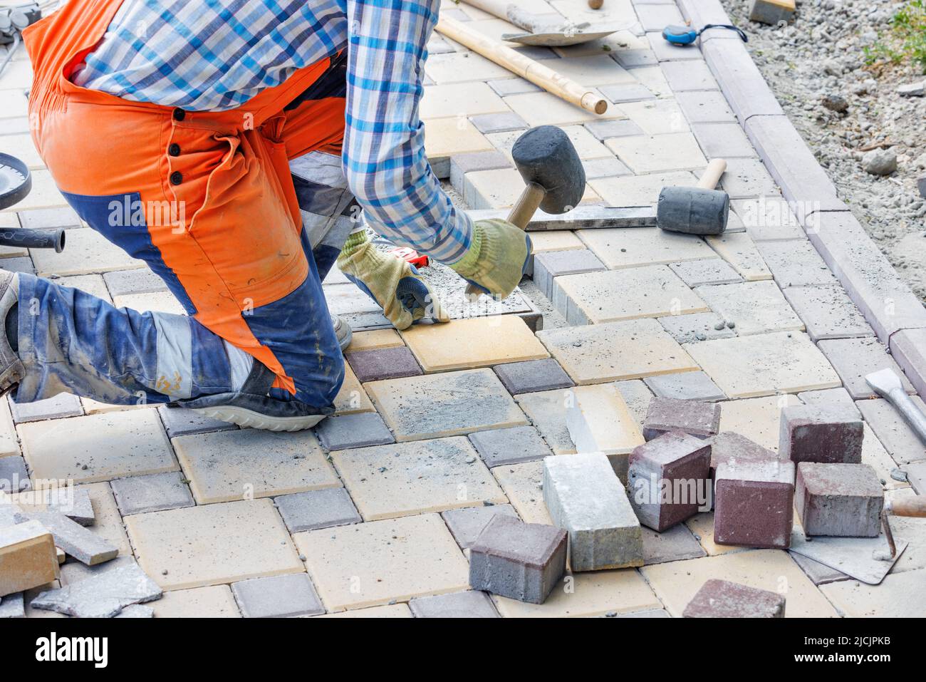A worker in blue and orange overalls squats and lays paving slabs with a rubber mallet. Closeup, selective focus, copy space. Stock Photo