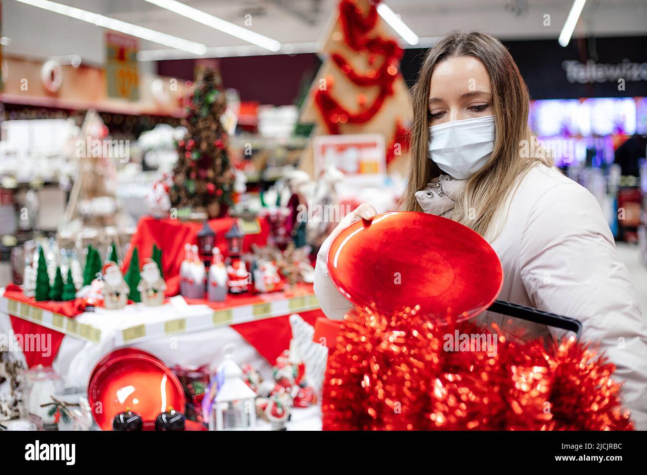 Woman in white winter jacket and protective mask in shopping mall chooses Christmas red decorations for home deco. Christmas shopping. Personal Stock Photo