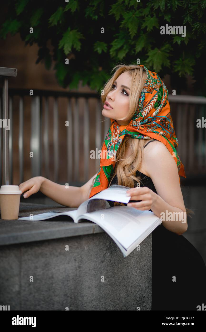young beautiful woman with headscarf on head is reading magazine on street, there is disposable cup of coffee, urban lifestyle Stock Photo