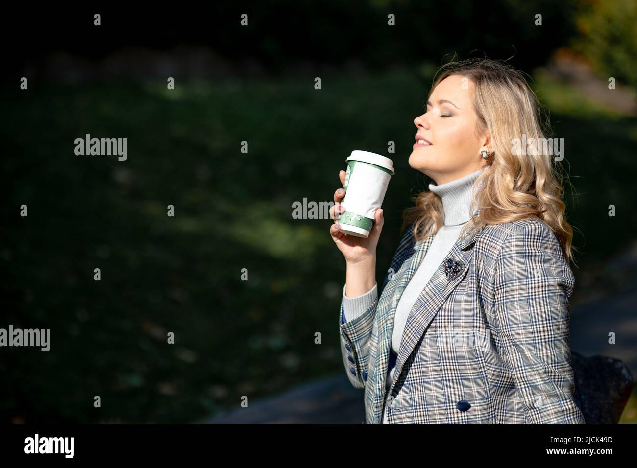 Attractive blonde with long curly hair in gray-blue tweed jacket and sweater, exposing her face to warm autumn sun, enjoys hot chocolate in park Stock Photo