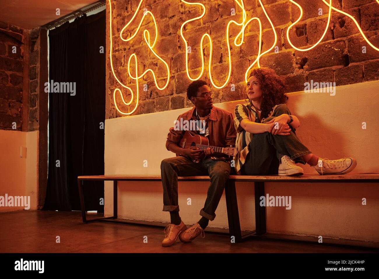 Young black man playing acoustic guitar while sitting on bench by brick wall next to his girlfriend in stylish casual apparel in studio Stock Photo