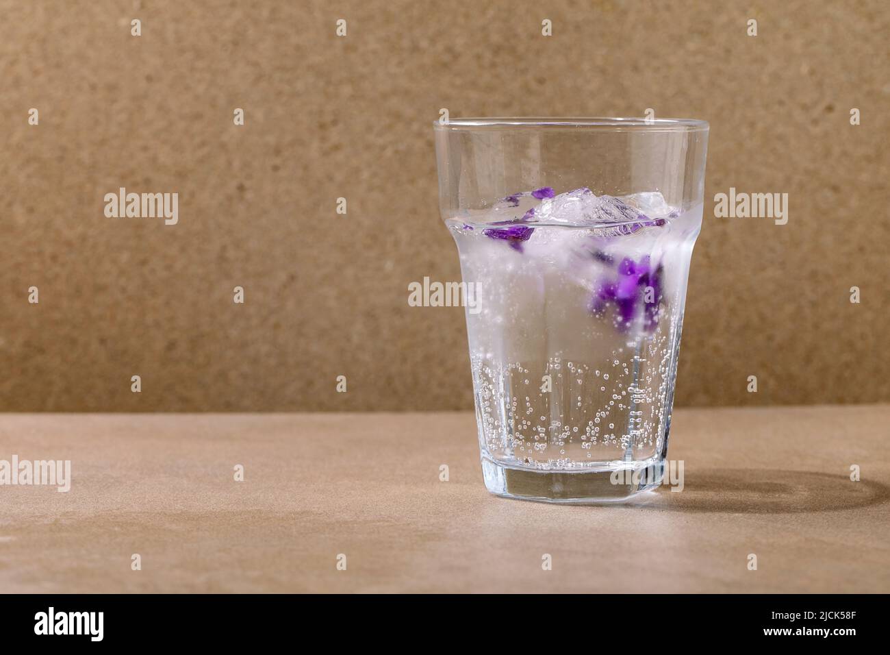 Transparent glass of sparkling soda with of violets flower ice cubes for making cold summer refreshment drink lemonade cocktail. Standing on brown tab Stock Photo