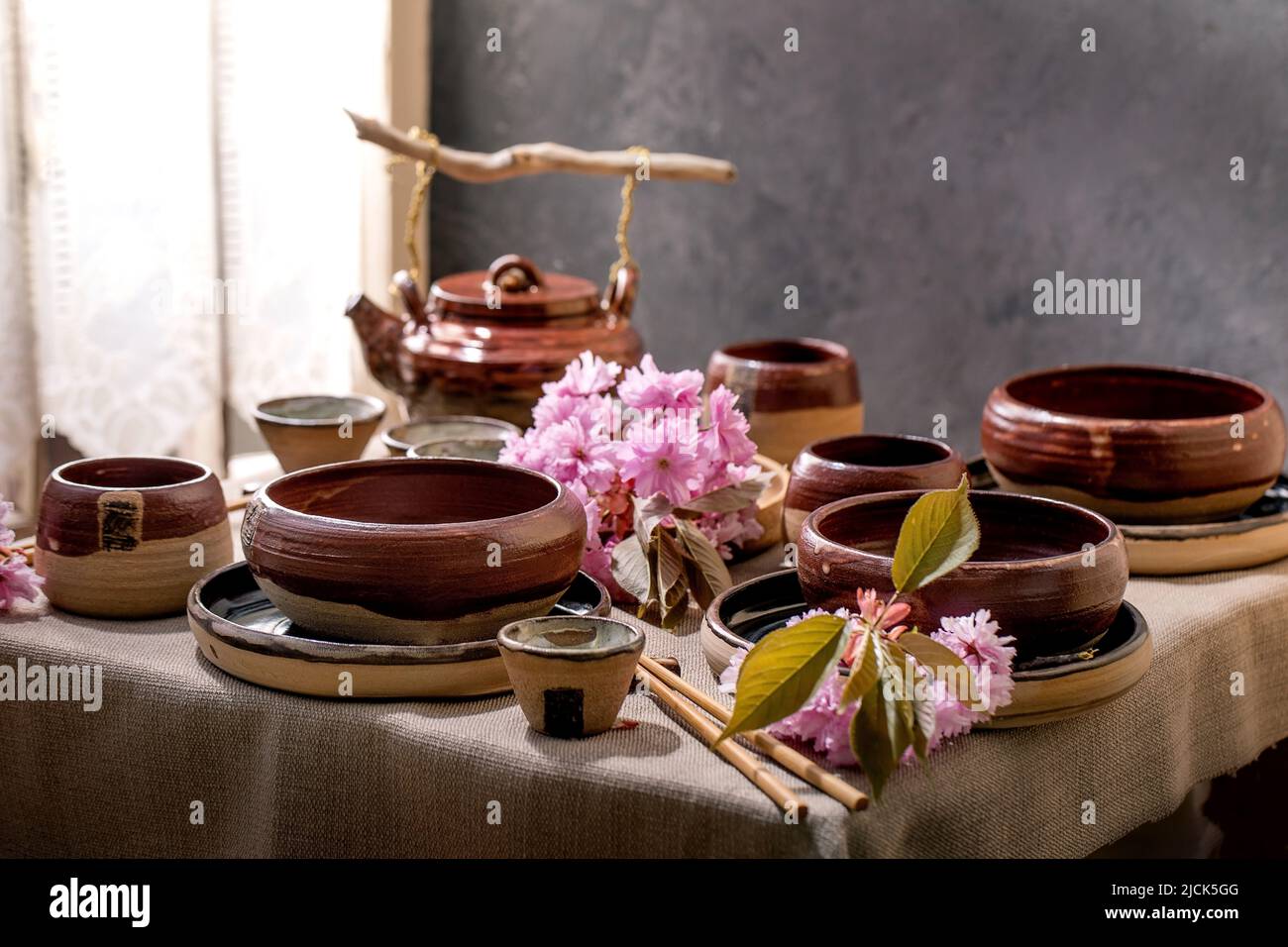 Japanese Asian style table setting with empty craft ceramic tableware, brown rough bowls, kettle and cups on linen tablecloth, decorated by pink sprin Stock Photo