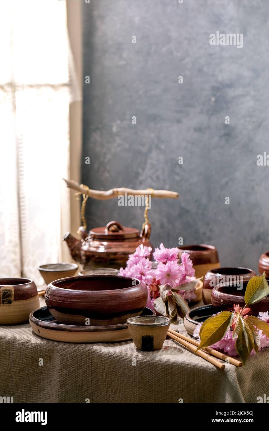 Japanese Asian style table setting with empty craft ceramic tableware, brown rough bowls, kettle and cups on linen tablecloth, decorated by pink sprin Stock Photo
