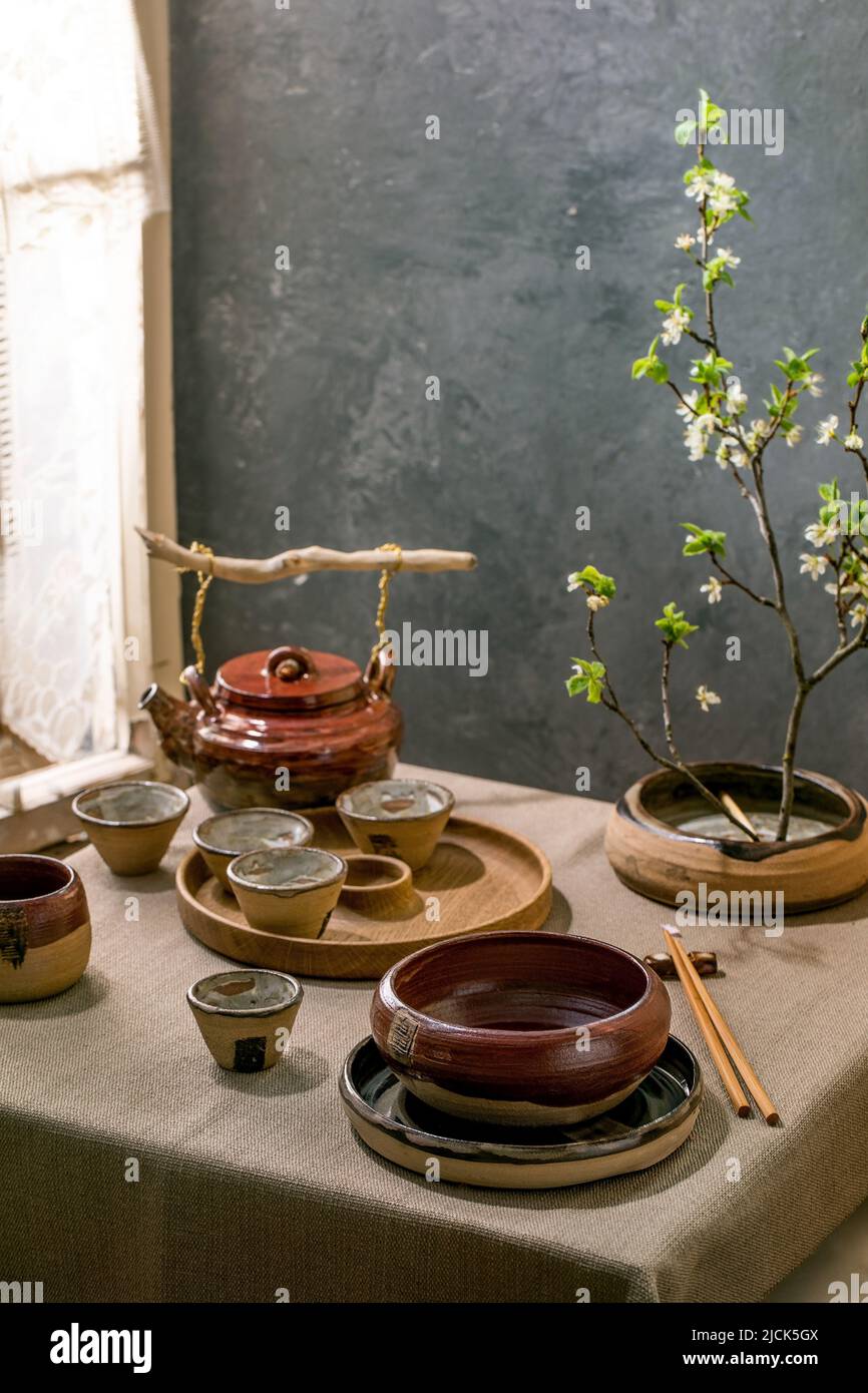 Japanese Asian style table setting with empty craft ceramic tableware, brown rough bowls and cups on linen tablecloth, decorated by ikebana spring blo Stock Photo