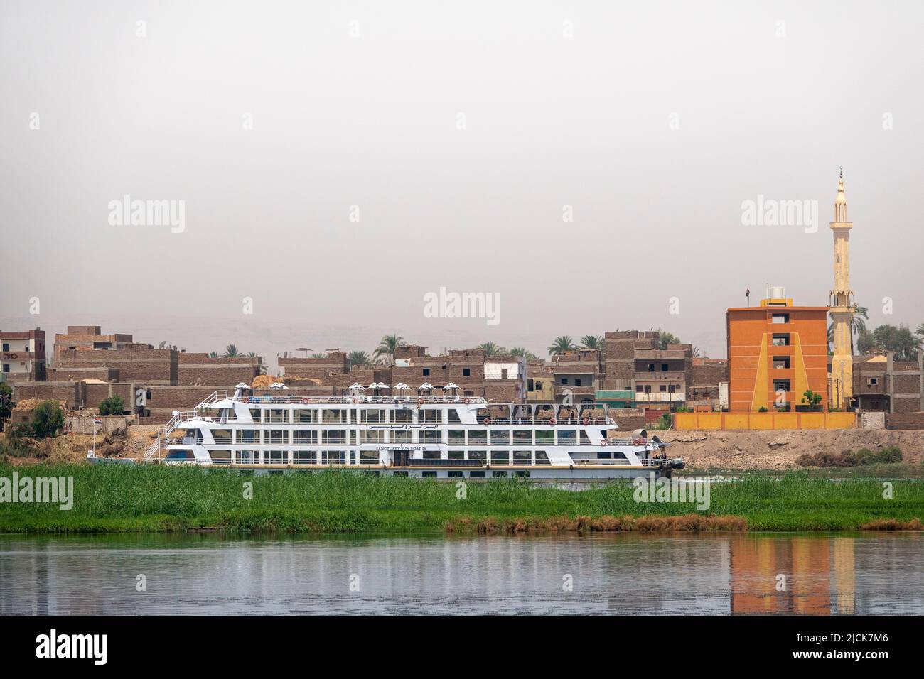 Nile cruise boat sailing side on to camera behind an island with band of grass with wall of buildings behind and band of river in foreground Stock Photo