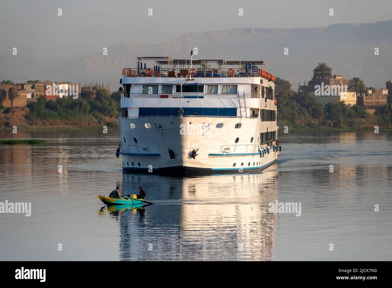 Nile cruise boat sailing on the calm water with reflections in early morning light heading straight towards a small two man fishing boat and camera Stock Photo