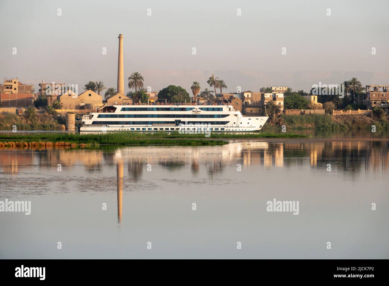 Nile cruise boat sailing side on to camera on the calm water with reflections of industrial scene behind in early morning light Stock Photo