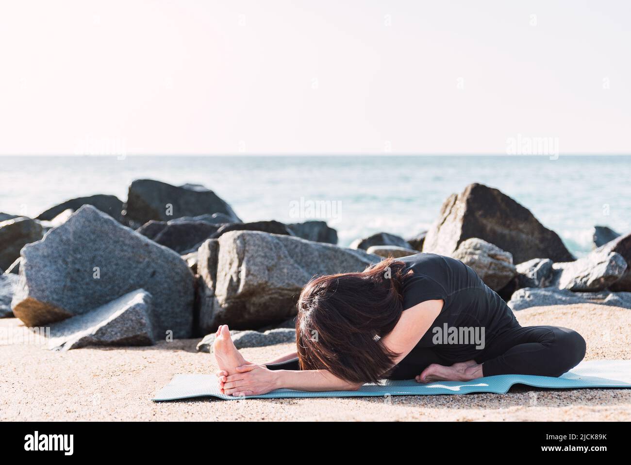 barefoot female in sportswear sitting on mat. She is in Head to Knee posture and stretching legs while doing yoga on shore near sea in sunny day and looking at camera Stock Photo