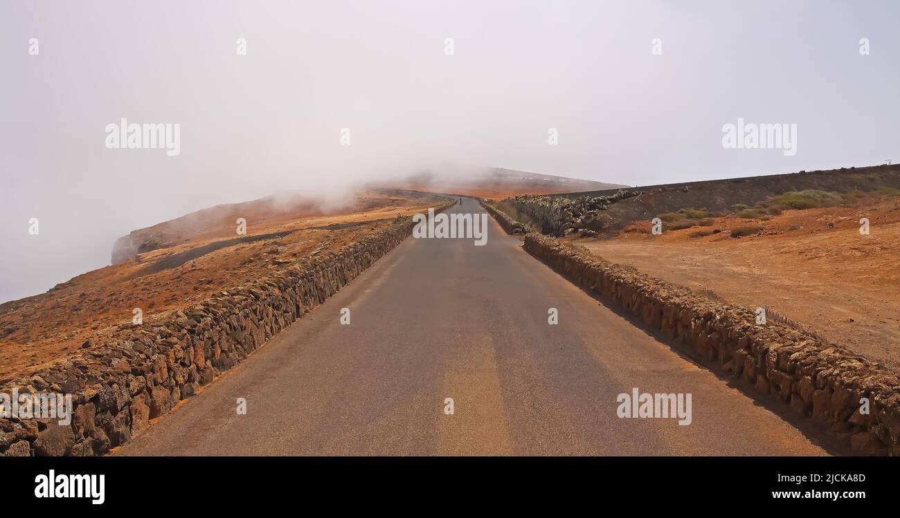 Scenic empty road on high mountain peak viewpoint leads into dense fog, natural stone wall close to abyss (Mirador del Rio - Lanzarote) Stock Photo