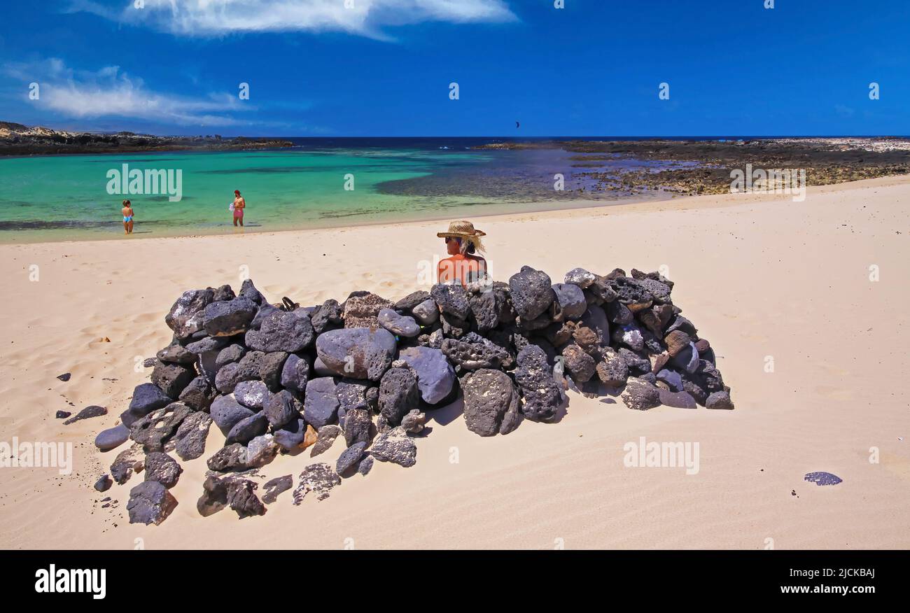 One blonde tourist woman sitting relax behind natural stone rocks wall wind shield, idyllic white sand beach, beautiful turquoise sea lagoon horizon, Stock Photo
