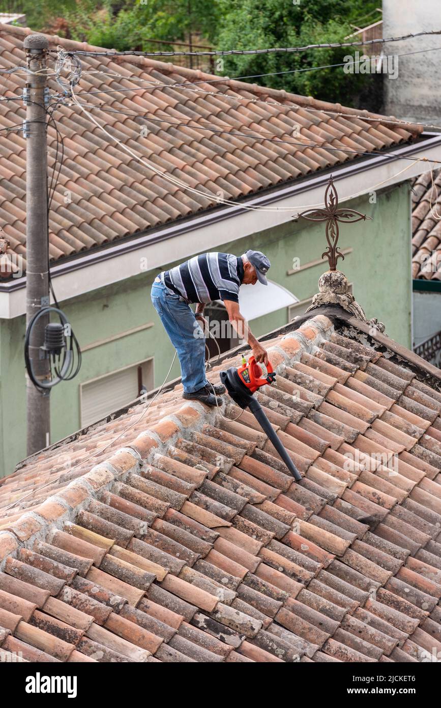 A man uses a leaf blower to remove volcanic ash from the roof of a chapel in the village of Milo, high on the slopes of Mount Etna, Sicily, Italy Stock Photo