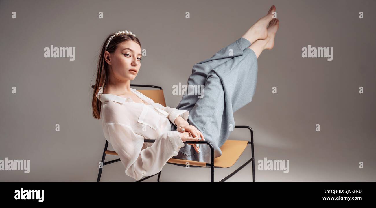 Casual young woman sits barefoot on a comfortable armchair, relaxes, raises her legs. Feet up.  Stock Photo