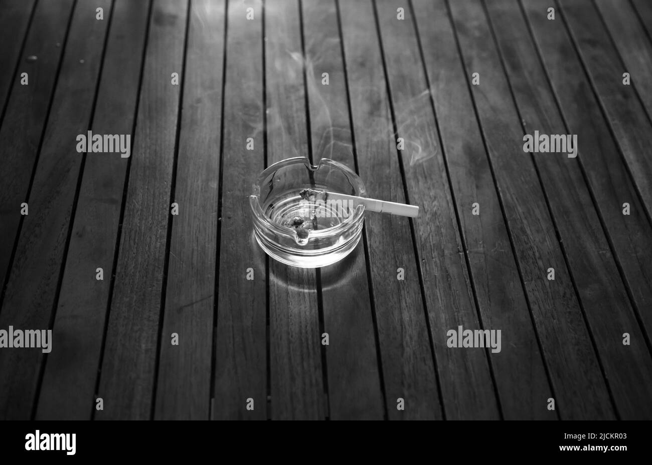Burning cigarette in a glass ashtray on a wooden table. Black and white. Stock Photo