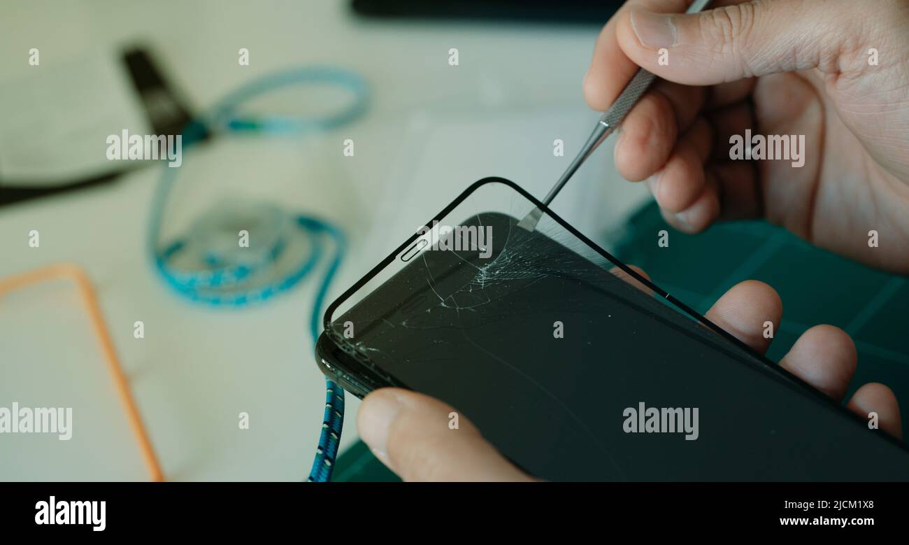 closeup a man, at a table, who is removing the broken screen protector of a smartphone using a metal utensil Stock Photo