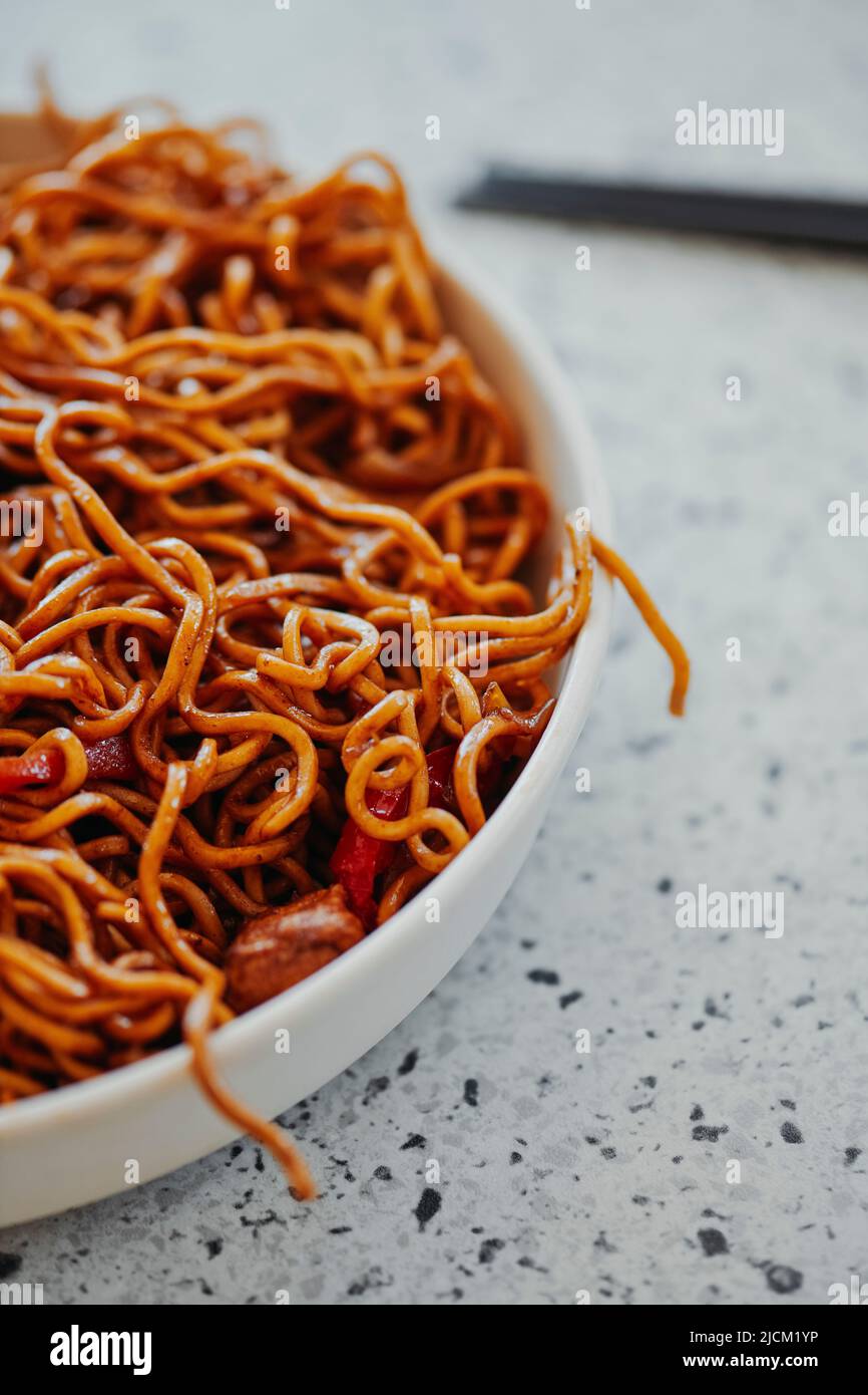 closeup of a plate with some chicken yakisoba noodles on a white mottled stone surface, next to a pair of black chopsticks Stock Photo
