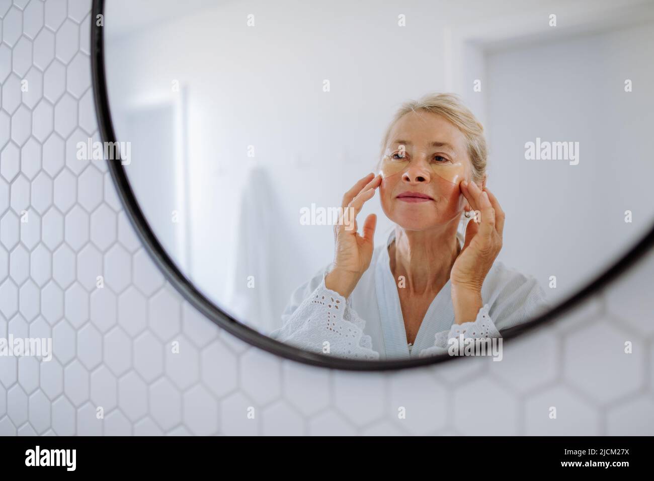 Beautiful senior woman in bathrobe, applying eye patches for puffiness while looking in the mirror Stock Photo