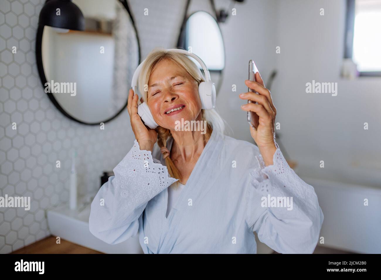 Beautiful senior woman in bathrobe listening to music in bathroom, relax and wellness concept. Stock Photo