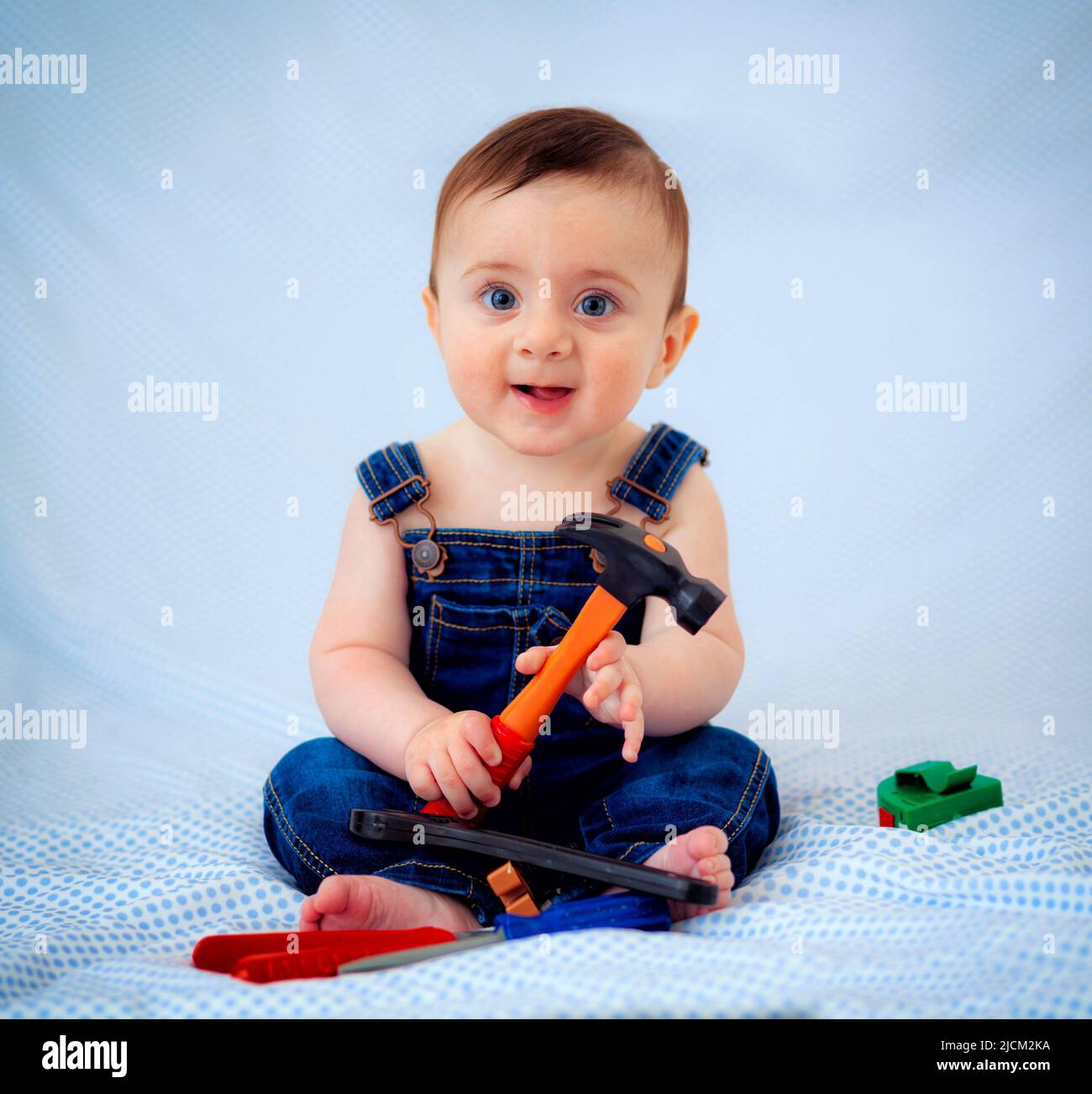 6-month-old baby boy dressed in denim overalls while playing tool worker. He looks like a little carpenter or a little plumber. Stock Photo
