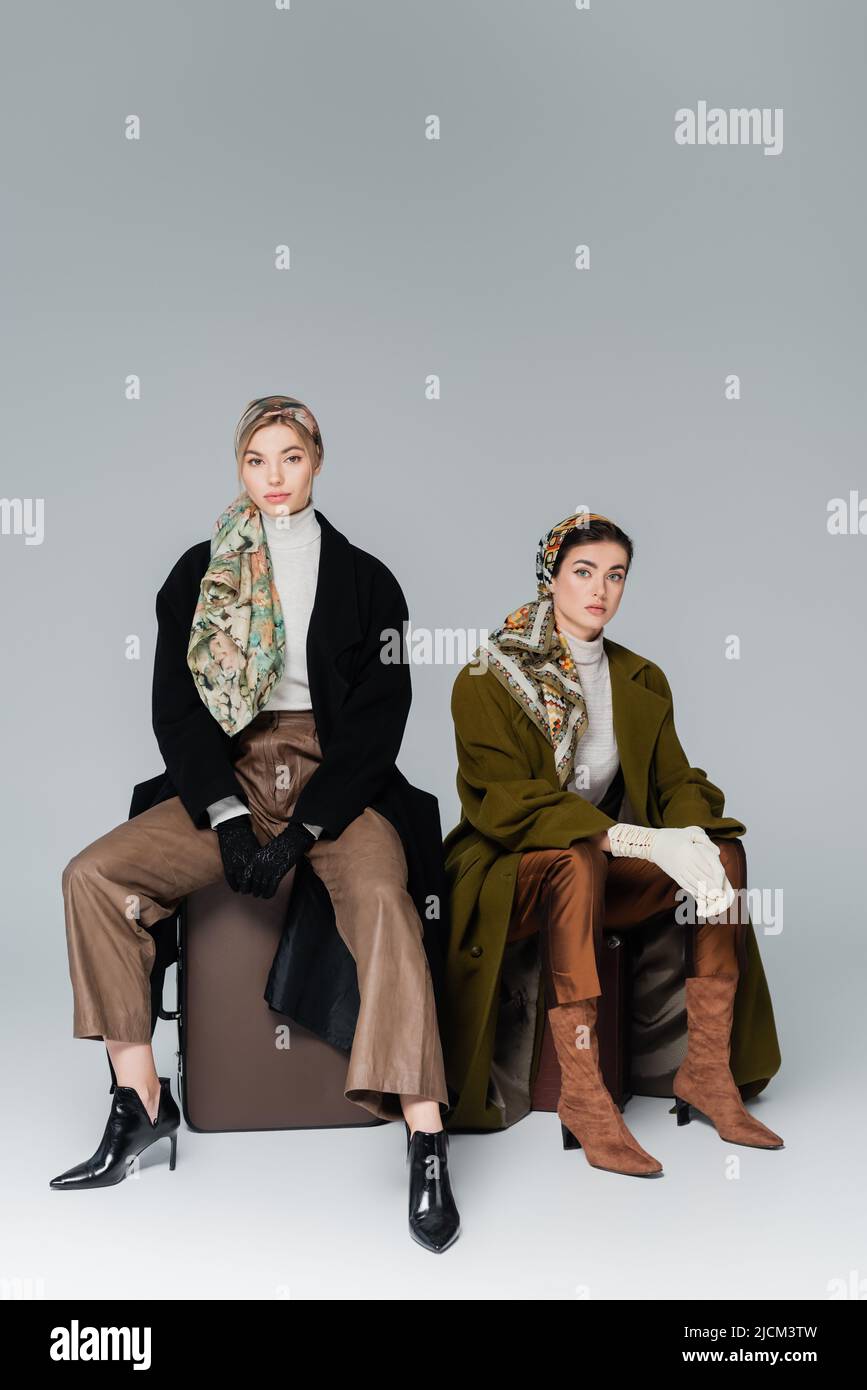 full length of fashionable women sitting on vintage suitcases on grey background Stock Photo