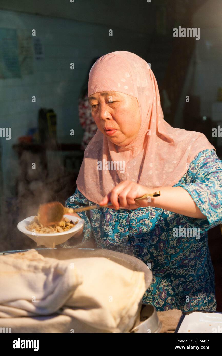 Middle aged women wearing Muslim head covering, in a local restaurant kitchen serving / serves hot steaming Chinese food into a dish to serve to a customer, to eat. Xi'an, China. (125) Stock Photo