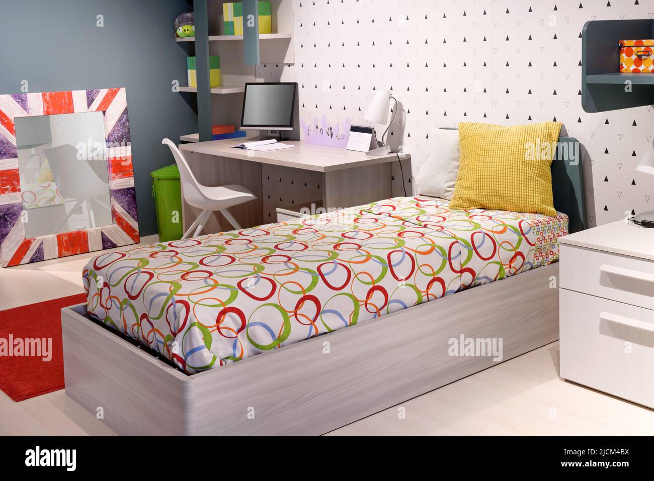 Comfortable bed with ornamental bedsheet and soft pillows located near desk with computer monitor and Union Jack mirror in modern teenager bedroom Stock Photo