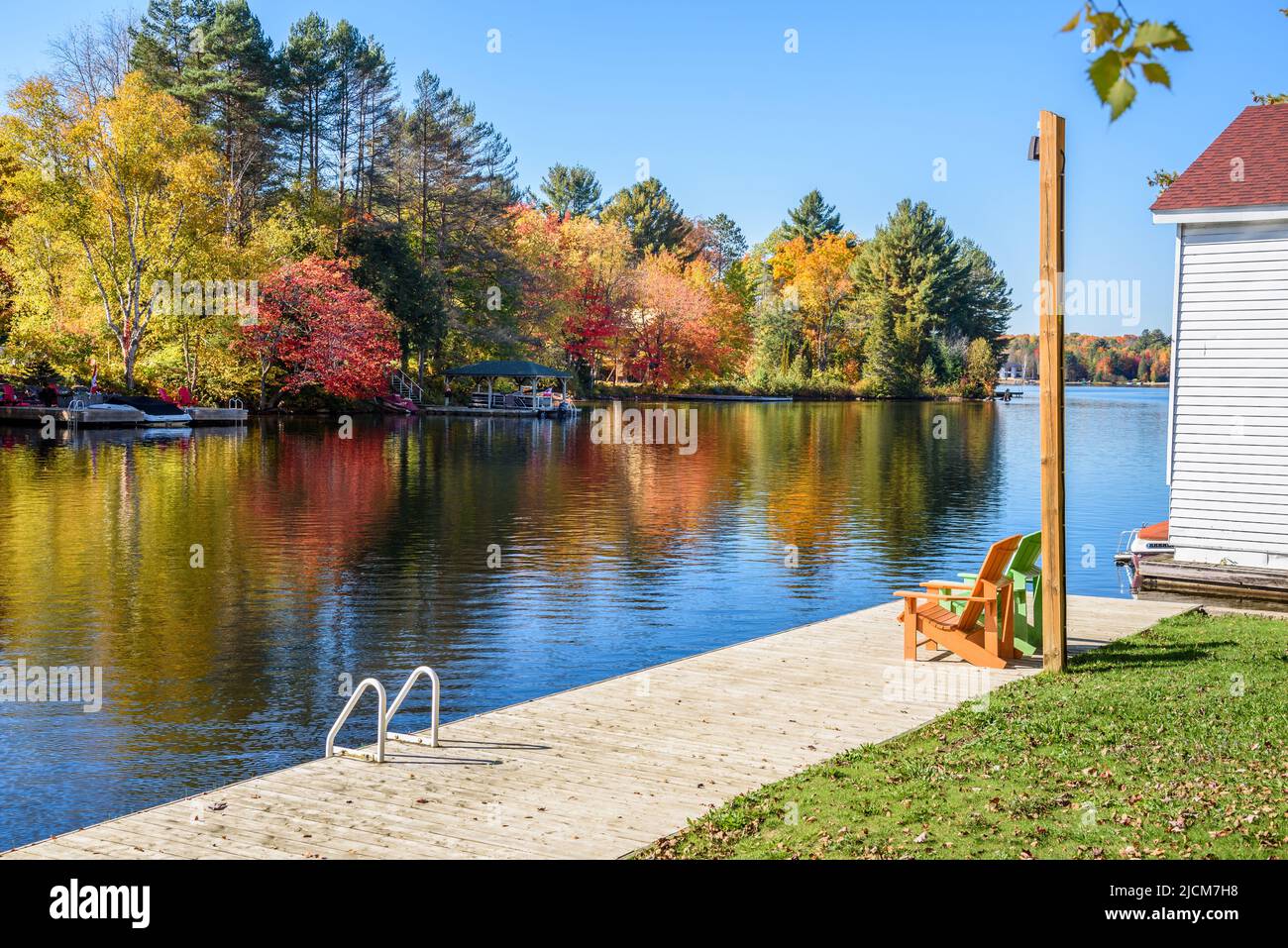 Deserted riverbank wooden boardwalk with two adirondack chairs on it on a sunny autumn day. Autumn colours reflectiong in water. Stock Photo