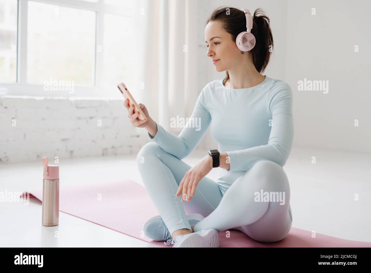 Sporty woman sitting on a yoga mat using smartphone and listening to music on headphones during training at gym. Stock Photo