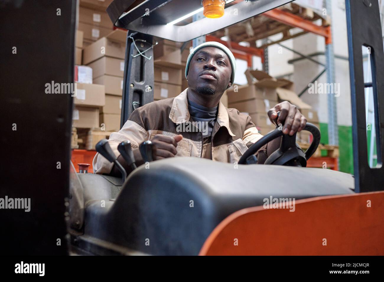 Serious African worker in uniform driving forklift at warehouse to load cargo Stock Photo