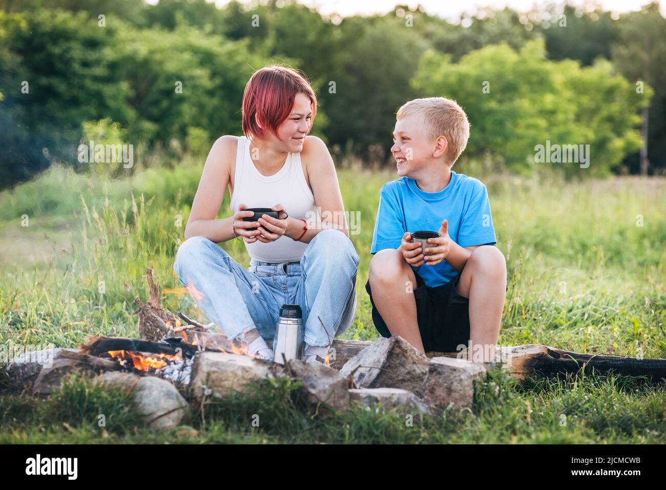 Smiling sister and brother kids have merry conversation near campfire. They looking eye to eye to each other, drinking tea from thermos. Happy family Stock Photo