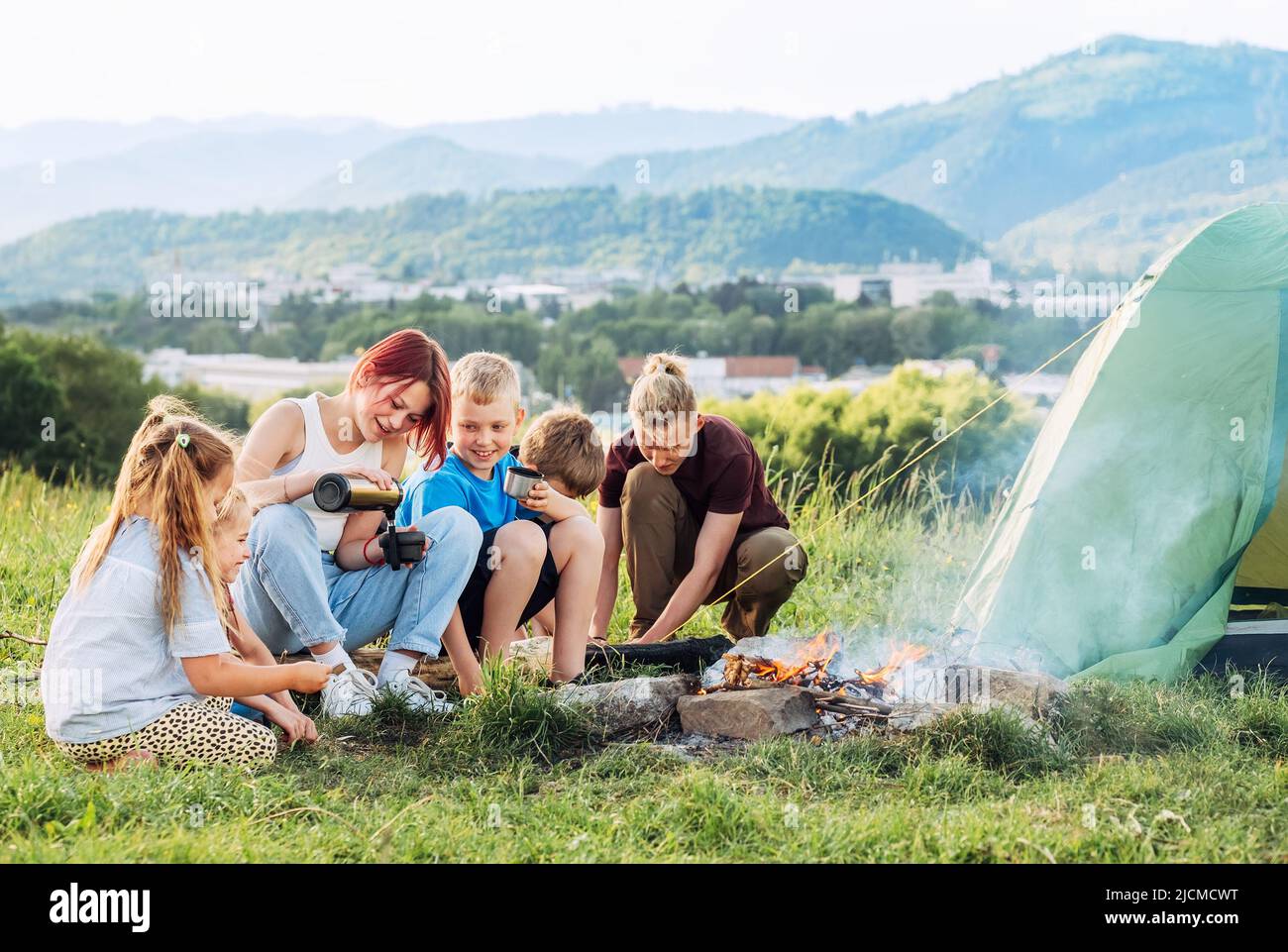 Group of smiling kids has a merry conversation near a smoky campfire. They drinking tea from a thermos, two brothers set up the green tent. Happy fami Stock Photo