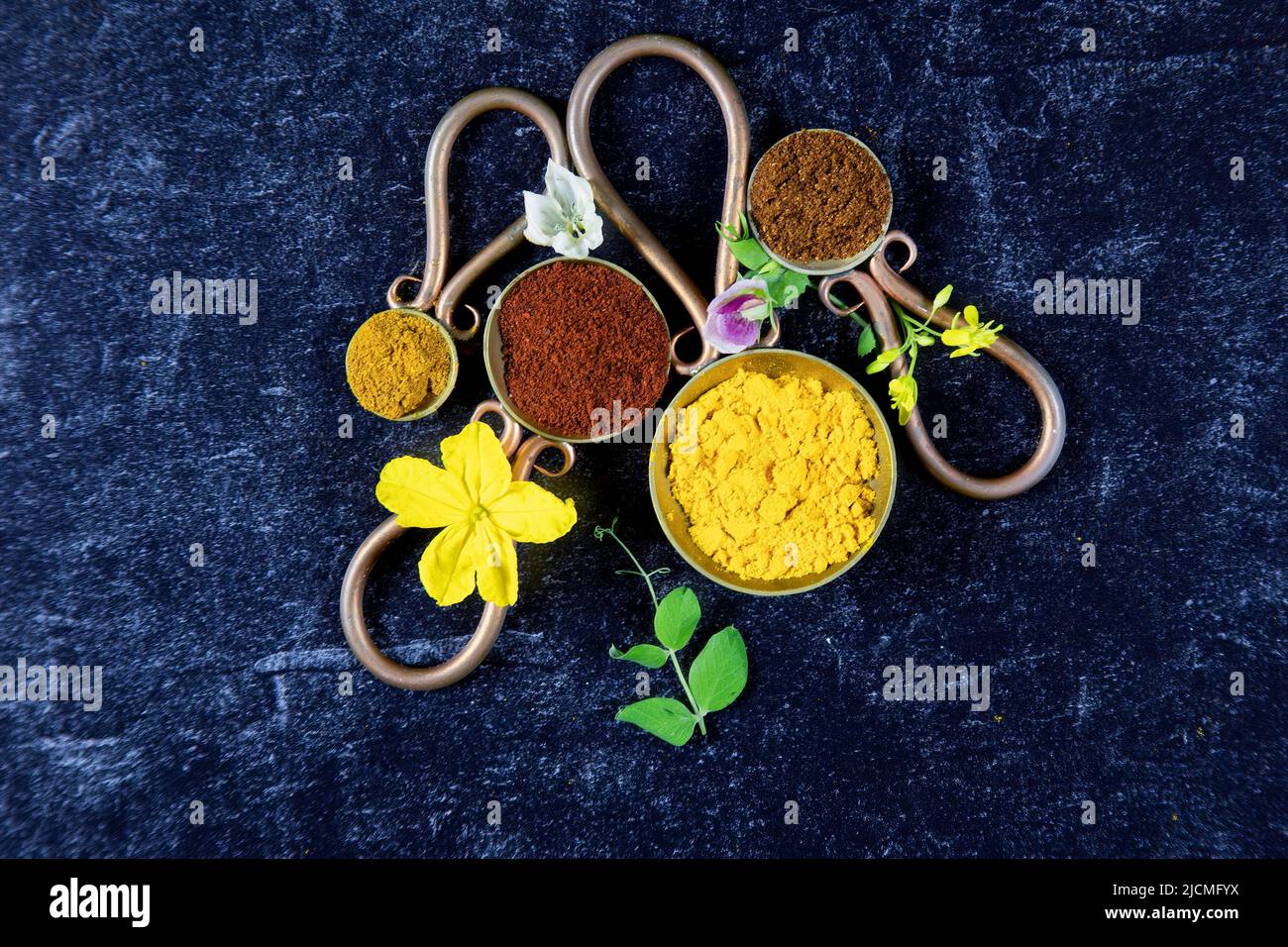Vintage metal spoons with ground spices of chili powder, tumeric, paprika and curry powder with fresh flowers from flowering vegetables on black with Stock Photo