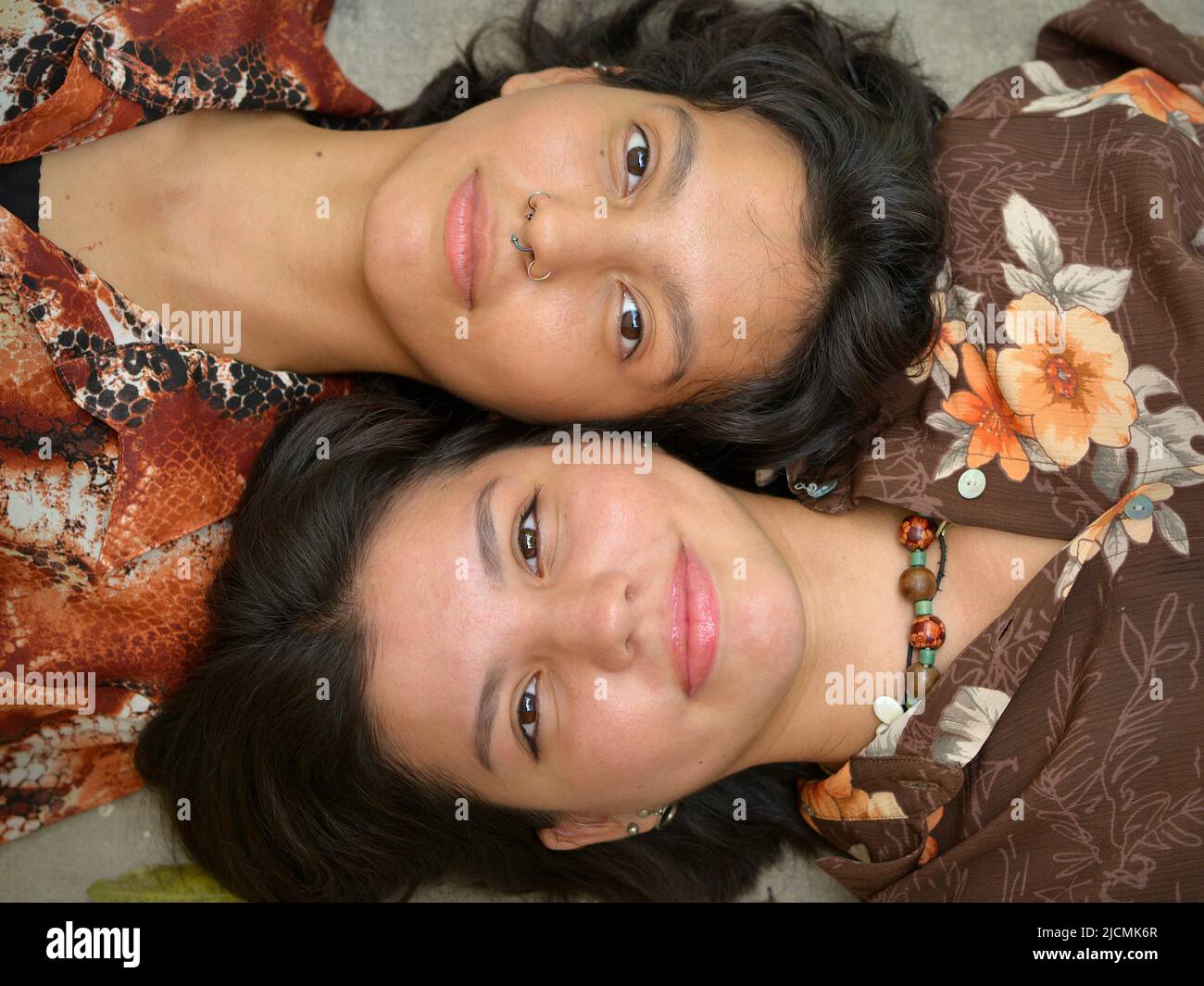 Two brown-eyed pretty attractive Mexican sisters (young adults) lie on the floor ground cheek to cheek side by side and look at the camera. Stock Photo