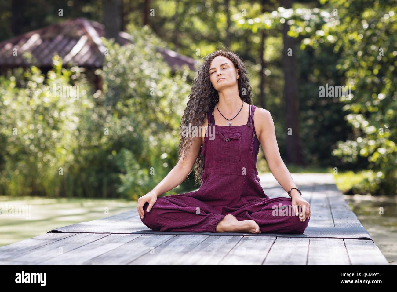 A woman practicing yoga, meditating with her eyes closed, sitting in a lotus position on a wooden bridge, on the shore of a pond on a warm sunny morni Stock Photo