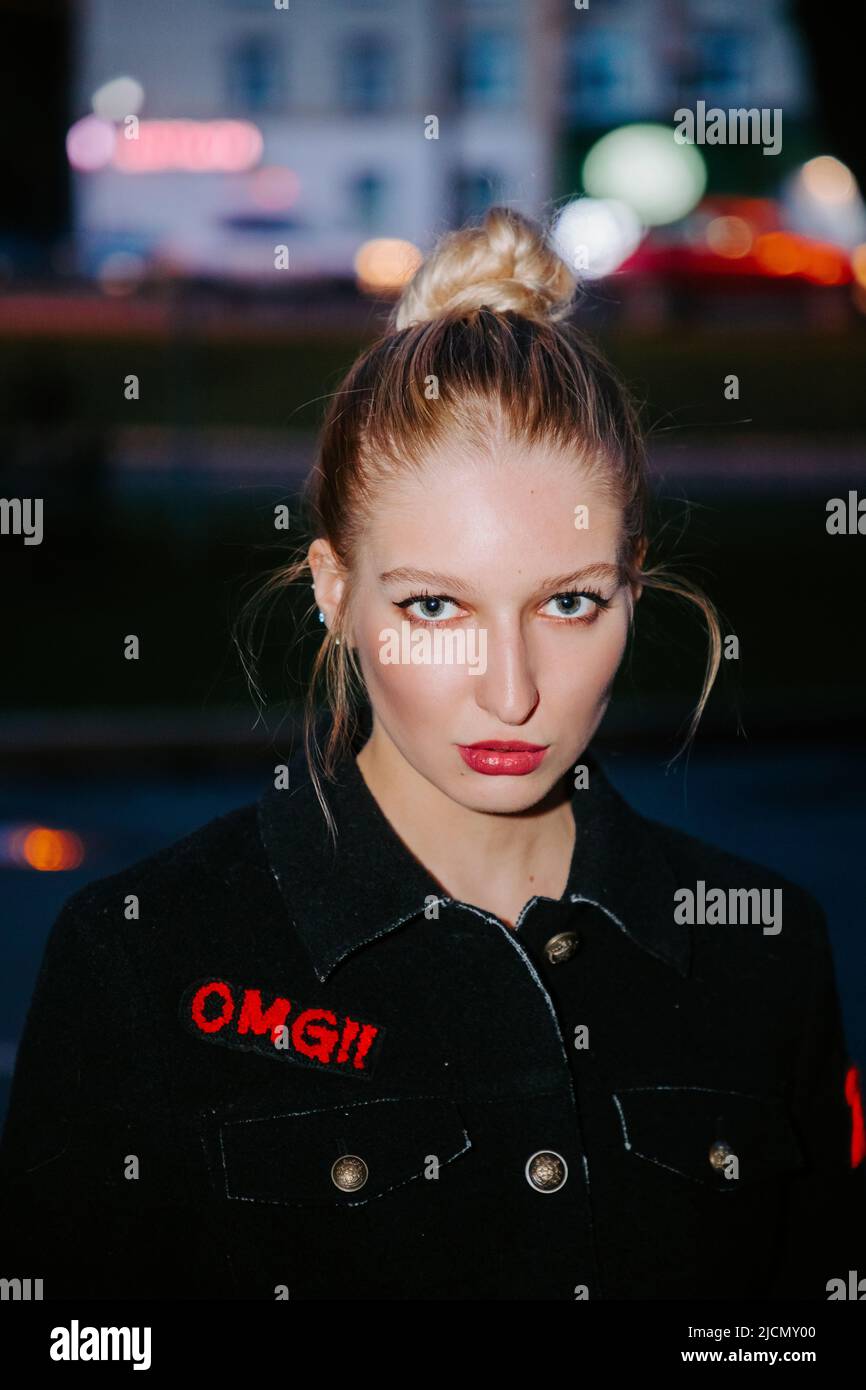 Portrait of a pretty blonde girl with red lips and a hair bun, wearing a black coat with 'OMG!' on it; she looks at us with intense eyes Stock Photo