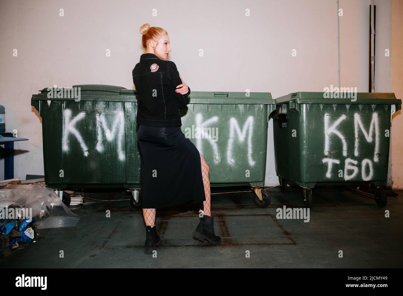 Pretty blonde girl with hair bun, wearing a long black coat and black boots, stands confidently near green waste containers with 'KM' painted on them Stock Photo