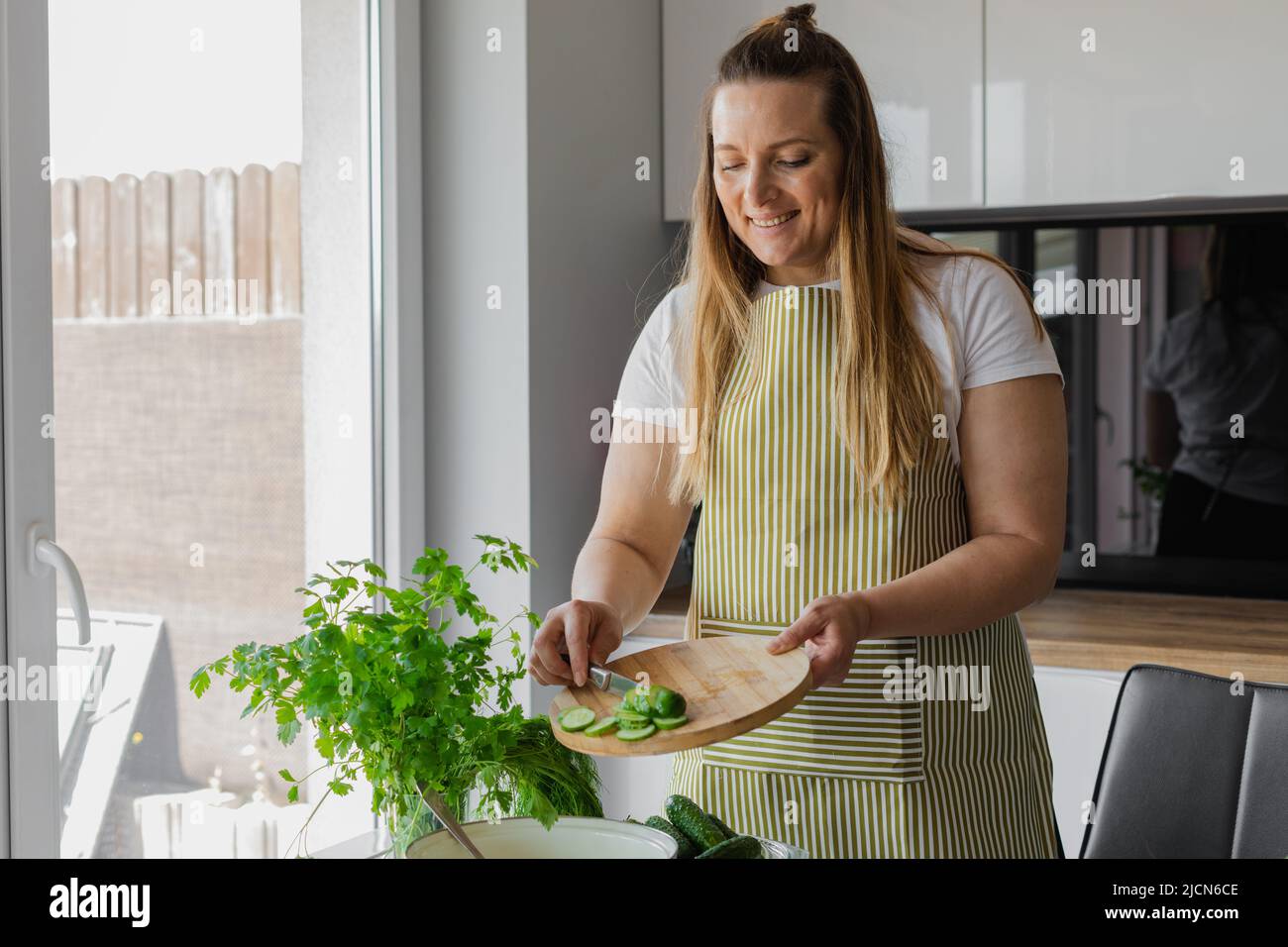 Smiling woman 40 years old in apron cutting cucumbers on cutting board in kitchen at home. Woman cooking Stock Photo